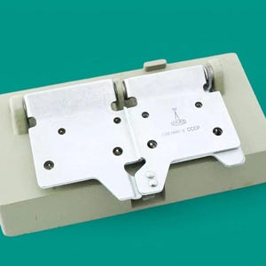 May include: A rectangular beige plastic base featuring a hinged metal mechanism with two silver plates. The right plate displays a small engraved icon of a radio tower and the text "СДЕЛАНО В СССР" in Cyrillic characters, indicating its origin.
