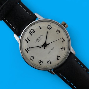 Puede incluir: Un reloj de pulsera vintage con caja plateada y esfera crema. El reloj presenta números arábigos y agujas negras, con la marca "Cardinal". Tiene una correa de cuero negro con costuras blancas, sobre un fondo azul.