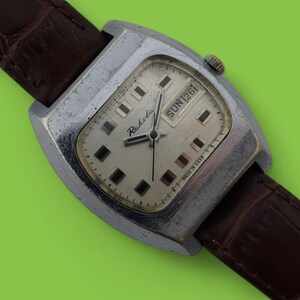 May include: Vintage Raketa watch with a silver-toned rectangular case and a light-colored face. The watch features a brown leather strap and displays the day of the week. The watch face has black square hour markers and black hands.