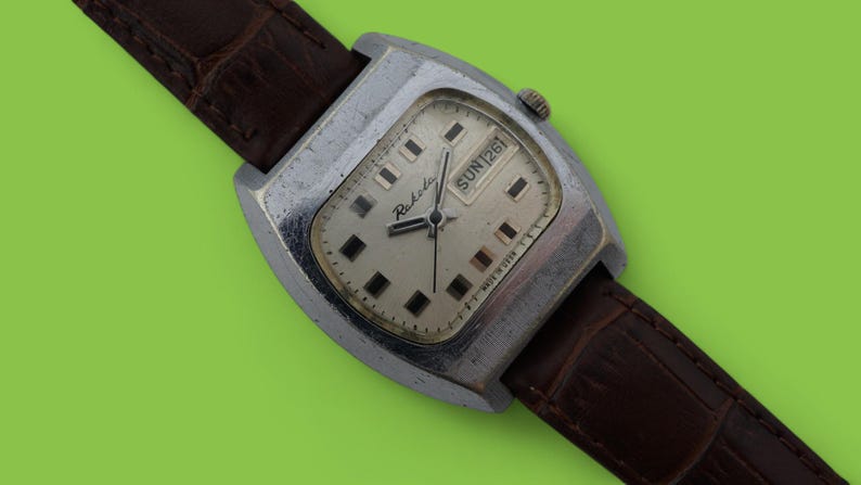 May include: Vintage Raketa watch with a silver-toned rectangular case and a light-colored face. The watch features a brown leather strap and displays the day of the week. The watch face has black square hour markers and black hands.