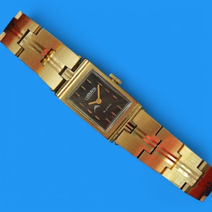 May include: A gold-toned wristwatch with a square face and a black dial. The watch has a gold-toned metal band with a unique design. The watch face has the text "CORNAVIN 17 Jewels" printed on it.