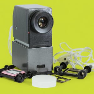 Könnte beinhalten: Ein Vintage-Diaprojektor mit schwarzem und silbernem Gehäuse, einem weißen Kabel und einem schwarz-weißen Diamagazin. Der Projektor steht auf einer gelben Oberfläche.