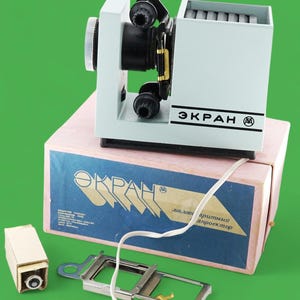Könnte beinhalten: Ein vintage Diaprojektor in hellblau mit einem weißen Kabel und einem weißen Stecker. Der Projektor ist mit "ЭКРАН" in schwarzen Buchstaben beschriftet. Der Projektor steht auf einem rosa Karton mit dem gleichen Label in blauen Buchstaben. Der Karton trägt außerdem die Aufschrift "для любительского кинопроектор".