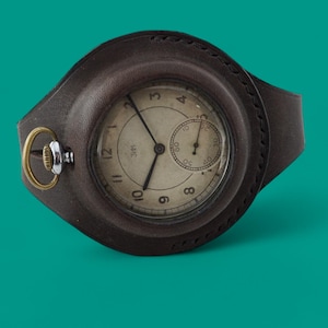 Puede incluir: Un reloj de pulsera de estilo vintage con una correa y una caja de cuero marrón. La esfera del reloj es blanquecina con números y agujas negras. Una esfera más pequeña está situada a las 3 en punto. El reloj tiene un anillo dorado y una corona plateada.