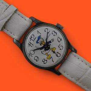 Puede incluir: Reloj de pulsera vintage con esfera blanca con una imagen de dibujos animados de Pato Donald. El reloj tiene manecillas y números negros, una caja plateada y una correa de cuero blanca. El reloj está sobre un fondo naranja.