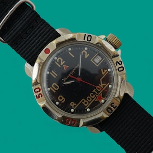 May include: Vintage Vostok watch with a black dial, red accents, and a black nylon strap. The watch has a gold-toned bezel with red numerals and a gold crown. The dial includes the word "Bostok" and a red star.