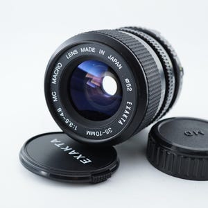 May include: A black Exakta 35-70mm camera lens with lens caps. The lens is marked "Made in Japan" and "MC Macro". The lens has a diameter of 52mm and an aperture of f/3.5-4.8.