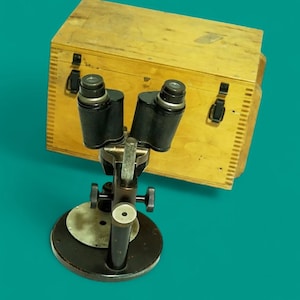Op de afbeelding: Een binoculaire microscoop met een zwart metalen frame en twee oculairs, gemonteerd op een ronde voet. Achter de microscoop staat een rechthoekige opbergdoos van lichtgekleurd hout met twee zwarte metalen sluitingen en zichtbare houtnerf.