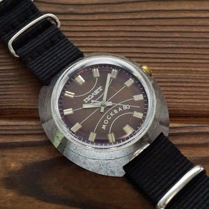 Pode incluir: Um rel&oacute;gio de pulso prateado com uma pulseira preta. O mostrador do rel&oacute;gio &eacute; de cor marrom escura com marcas prateadas e as palavras "POABET" e "MOCKBA 80".