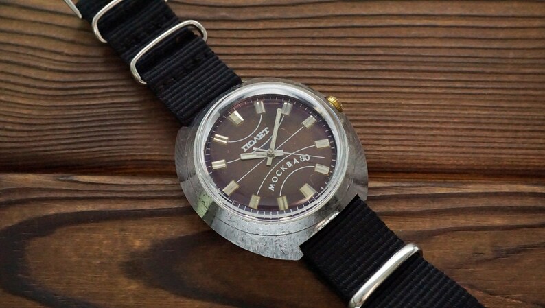 Pode incluir: Um rel&oacute;gio de pulso prateado com uma pulseira preta. O mostrador do rel&oacute;gio &eacute; de cor marrom escura com marcas prateadas e as palavras "POABET" e "MOCKBA 80".