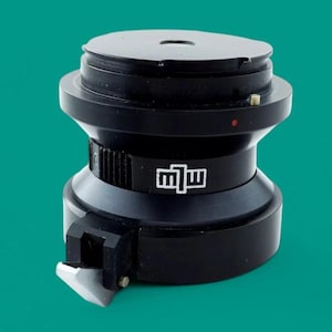 May include: A black cylindrical mechanical component featuring a stepped design with a flat top surface. The side displays a white logo with the letters "mlw" and a small red dot indicator. A silver-colored metal blade attachment is fixed to the base.