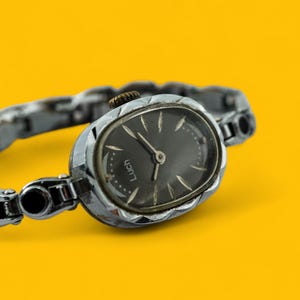 May include: A silver wristwatch with a black face and a metal band. The watch face has a black background with white hour and minute hands and a silver second hand. The watch face also has the word "LUCH" printed in white.