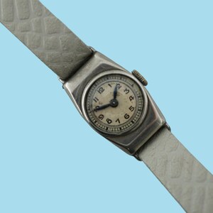 Könnte beinhalten: Vintage silberfarbene Armbanduhr mit hellem Zifferblatt und schwarzen Zeigern. Die Uhr hat ein rechteckiges Gehäuse und ein hellgraues, strukturiertes Lederarmband. Das Zifferblatt zeigt Zahlen und Stundenmarkierungen.