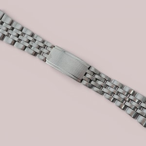 May include: A silver-toned metal watch band featuring a multi-link design with a central rectangular clasp. The clasp has a subtle horizontal ribbed texture, and the band consists of several rows of small, polished metallic links connected together.