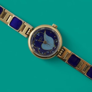 Puede incluir: Un reloj de pulsera dorado con esfera azul y correa dorada y azul. La esfera del reloj presenta un diseño de hoja azul claro. La correa tiene detalles rectangulares azules. El reloj está sobre un fondo turquesa.