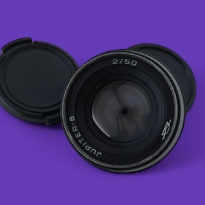 Puede incluir: Un objetivo de cámara negro Jupiter-8 2/50 con una tapa de objetivo a juego, sobre un fondo morado. El objetivo tiene letras blancas que dicen "2/50" y "Jupiter-8". La tapa del objetivo es redonda y negra.