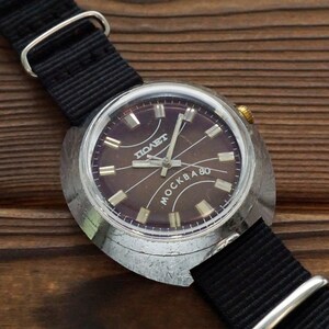 Pode incluir: Um rel&oacute;gio de pulso prateado com uma pulseira de couro preta. O mostrador do rel&oacute;gio &eacute; de cor marrom escura com marcas prateadas e as palavras "ПОЛЕТ" e "MOCKBA 80".