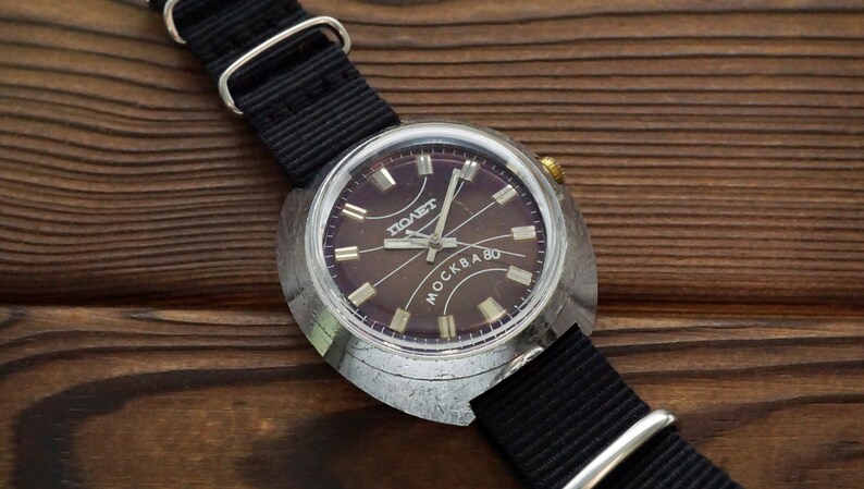 Pode incluir: Um rel&oacute;gio de pulso prateado com uma pulseira de couro preta. O mostrador do rel&oacute;gio &eacute; de cor marrom escura com marcas prateadas e as palavras "ПОЛЕТ" e "MOCKBA 80".