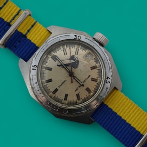 Op de afbeelding: Een vintage polshorloge met een zilverkleurige kast en een crèmekleurige wijzerplaat. Het horloge heeft een blauw-gele gestreepte nylon band. De wijzerplaat toont de cijfers 15, 30 en 45. Het horloge staat tegen een turquoise achtergrond.