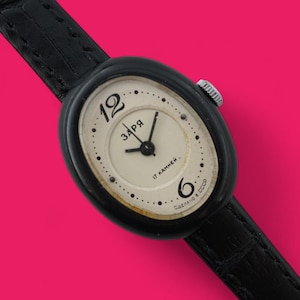 May include: Vintage black wristwatch with an oval face and a black leather band. The watch face is cream-colored with black numerals and hands. The text "ЗАРЯ" is visible on the watch face. The watch is set against a bright pink background.