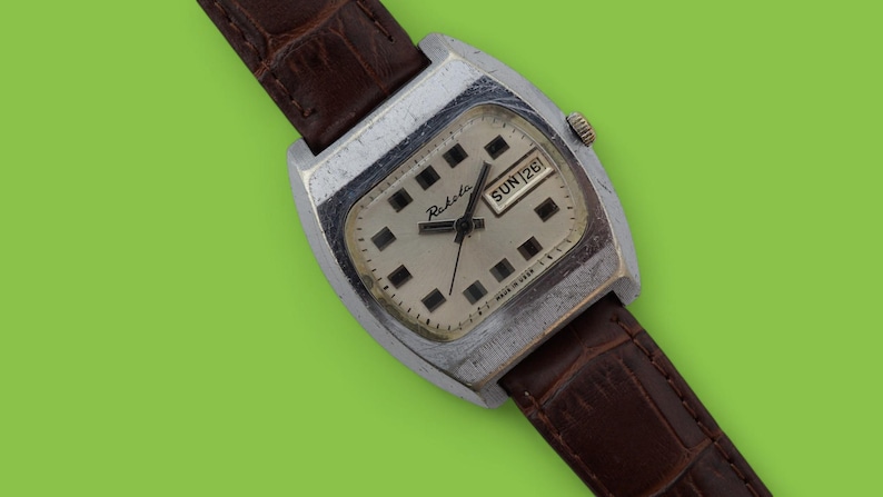 May include: Vintage Raketa watch with a silver-tone case and a brown leather band. The watch face is off-white with black square hour markers and the day and date displayed. The watch has a textured brown leather band.