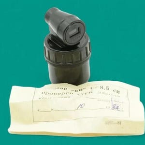 Puede incluir: Un componente óptico cilíndrico negro con un visor montado en la parte superior, acompañado de un documento de papel rectangular de color blanco roto con texto en cirílico, la designación F=8,5 cm y una fecha manuscrita de 1982.