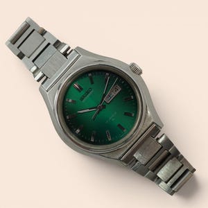 May include: A silver-toned Seiko wristwatch featuring a vibrant emerald green circular dial with silver hour markers and hands. The watch includes a day-date window at the three o'clock position and a matching silver-toned metal link bracelet.