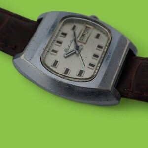 May include: A vintage wristwatch with a silver-colored, rectangular case and a light-colored face. The watch has a brown leather band. The watch face displays the day of the week and the time. The watch is set against a green background.