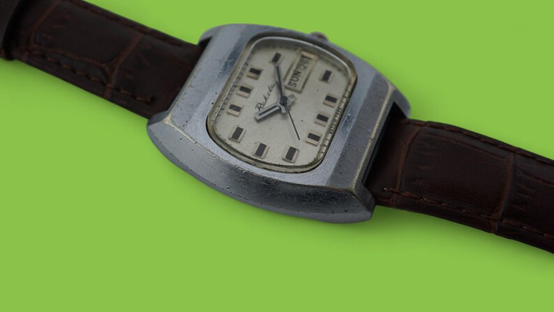 May include: A vintage wristwatch with a silver-colored, rectangular case and a light-colored face. The watch has a brown leather band. The watch face displays the day of the week and the time. The watch is set against a green background.