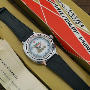 Può includere: Un orologio da polso nero e argento con quadrante bianco e bandiere rosso, bianco e blu. L'orologio è in una scatola con la scritta "Vostok Watch Maker Military Watch" su di essa.