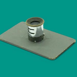 Puede incluir: Una placa rectangular de metal gris con una lupa cilíndrica montada en el centro. La lupa tiene un cuerpo de tono plateado pulido con un borde texturizado de color latón en la parte superior y una pequeña ranura de ajuste lateral.