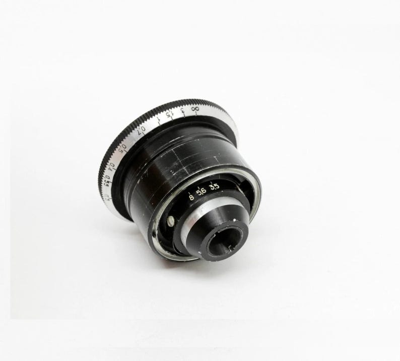 K&ouml;nnte beinhalten: Ein schwarzes Kameraobjektiv mit einem silbernen Einstellring. Das Objektiv hat eine Reihe von Zahlen und Markierungen auf dem Ring, darunter 8, 5,6 und 3,5. Das Objektiv ist leicht angewinkelt und gibt den Blick auf seine inneren Komponenten frei.