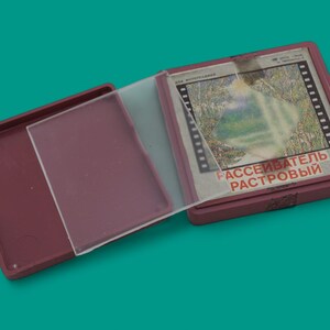 May include: A maroon plastic case, open to reveal a clear plastic sheet and a film-like image. The image is in a maroon frame and has text in Russian. The case is on a teal background.