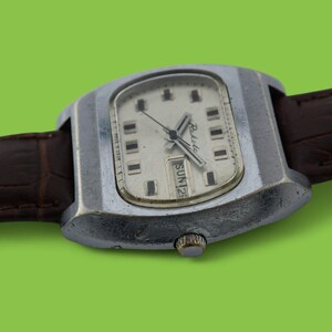 May include: Vintage wristwatch with a silver-toned case and a cream-colored face. The watch features black hour markers and hands, with a day and date display. It has a brown leather strap and a silver-toned crown. The watch is set against a green background.