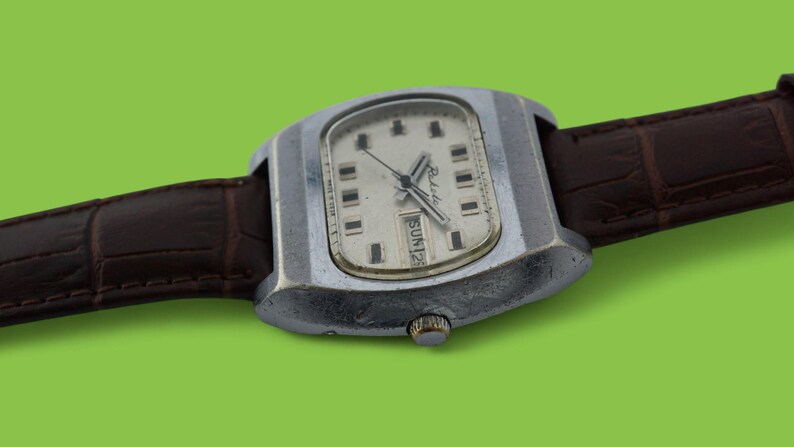 May include: Vintage wristwatch with a silver-toned case and a cream-colored face. The watch features black hour markers and hands, with a day and date display. It has a brown leather strap and a silver-toned crown. The watch is set against a green background.