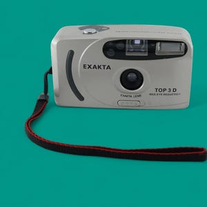 Könnte beinhalten: Cremefarbene Exakta Top 3D Kamera mit schwarzem und rotem Handgelenkband. Die Kamera hat einen Blitz, ein Objektiv und den Text "EXAKTA" und "TOP 3 D". Der Hintergrund ist türkis.