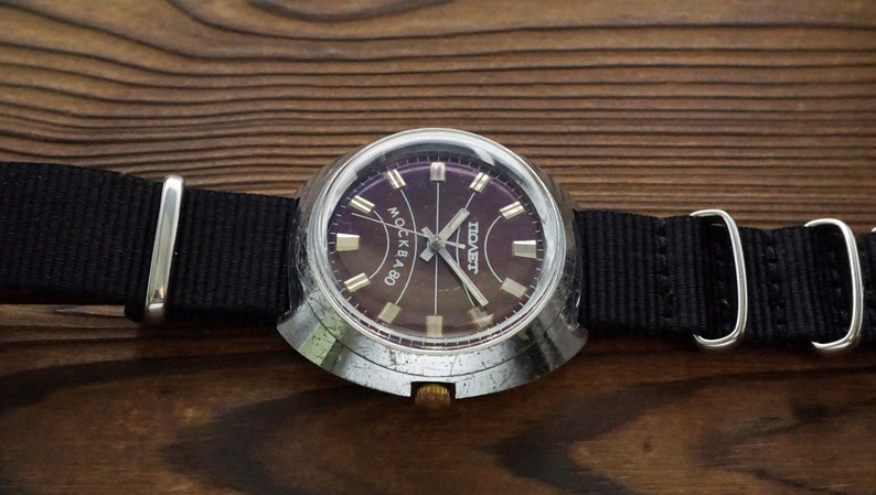 Pode incluir: Um rel&oacute;gio de pulso prateado com uma pulseira de nylon preta. O mostrador do rel&oacute;gio &eacute; roxo escuro com ponteiros e marcadores prateados. O mostrador do rel&oacute;gio tamb&eacute;m tem o texto "МОСКВА" e "СССР" impresso nele.