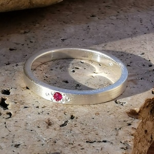 Puede incluir: Un anillo de banda de plata con una sola piedra roja engastada en el centro.