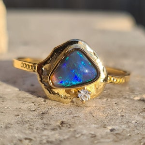 Peut inclure: Une bague en or ornée d'une opale triangulaire irisée, affichant des reflets bleus et verts. Un petit diamant est serti près de l'opale. La bague présente un design texturé et organique, évoquant une forme naturelle.