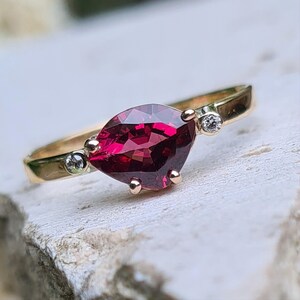 Op de afbeelding: Een gouden ring met een opvallende, dieprode marquise-geslepen edelsteen. De ring heeft twee kleine, heldere, ronde edelstenen. De band is van gepolijst goud. Een sieraad.