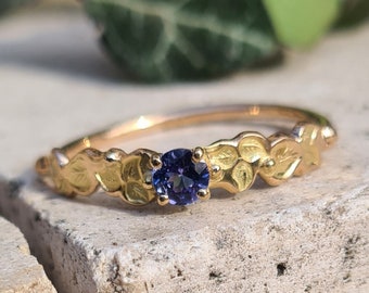 May include: A gold ring with a purple gemstone set in the center. The band is decorated with a row of leaves.
