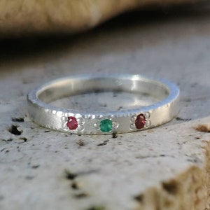 May include: A silver band ring with three small gemstones. The ring has a textured surface and features a red, green, and red gemstone.