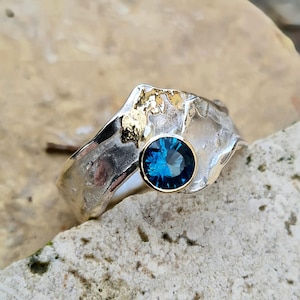 Peut inclure: Bague en argent avec un design texturé et un accent doré. La bague présente une pierre précieuse bleue foncée, ronde et facettée, sertie dans une lunette dorée. La bague est présentée sur une surface en pierre claire.