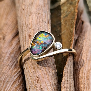 Pode incluir: Um anel de ouro com uma opala negra em forma de lágrima com flashes multicoloridos vibrantes de vermelho, verde e amarelo, acentuado por um pequeno diamante branco redondo cravado numa banda de estilo bypass que contorna as pedras.