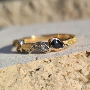 Peut inclure: Une bague en or avec une bande texturée et un design délicat. La bague est ornée d'une pierre précieuse noire en forme de poire et d'une petite pierre blanche.