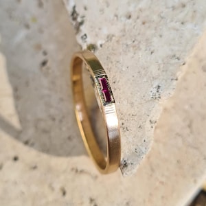 Op de afbeelding: Een gouden ring met een enkele, kleine, rechthoekige robijn die in het midden is gezet.