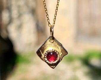 May include: A gold necklace with a small, red gemstone set in a gold pendant shaped like a diamond. The pendant has a textured surface.