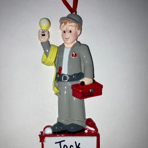 May include: A Christmas ornament depicting a cartoon electrician holding a lightbulb. The figure wears a gray uniform, a tool belt, and a cap. A red toolbox with "2020" is in his hand. The ornament is labeled "Jack" on a white base.