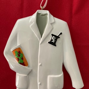 Puede incluir: Adorno de cerámica blanca con forma de bata de médico con un bolsillo en el lado izquierdo y un símbolo de farmacia en el lado derecho.