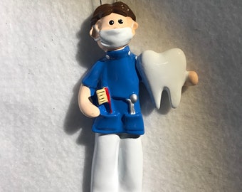 May include: A blue and white ceramic ornament of a dentist wearing a face mask and holding a large white tooth. The dentist is holding a toothbrush and a dental tool in their other hand.
